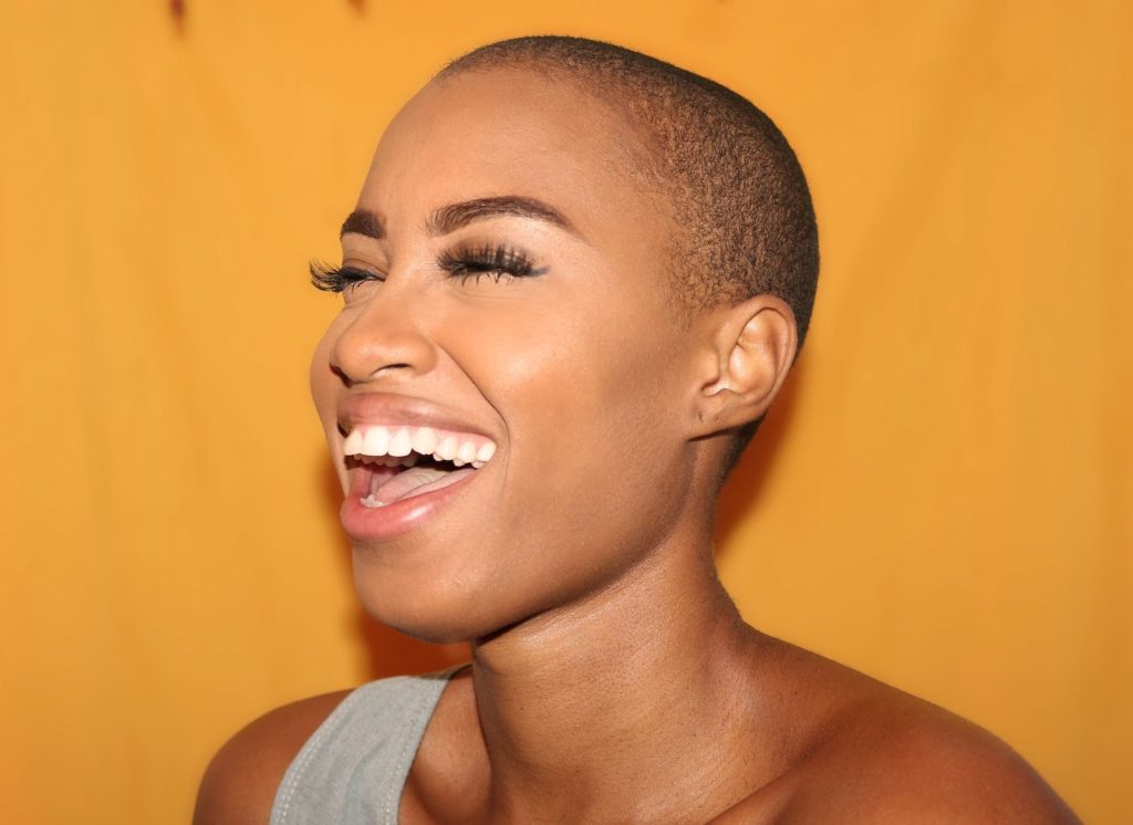 A person with a shaved head laughing against a warm yellow background, showcasing joy and positivity