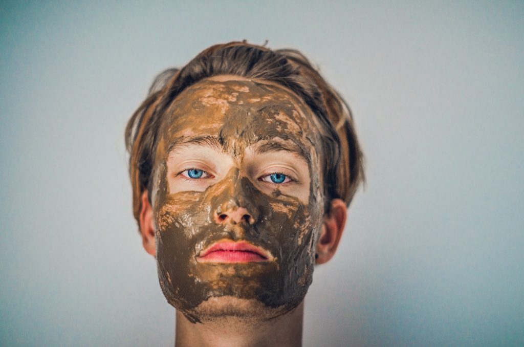 Man with mud facial mask for skincare and relaxation therapy against a plain background