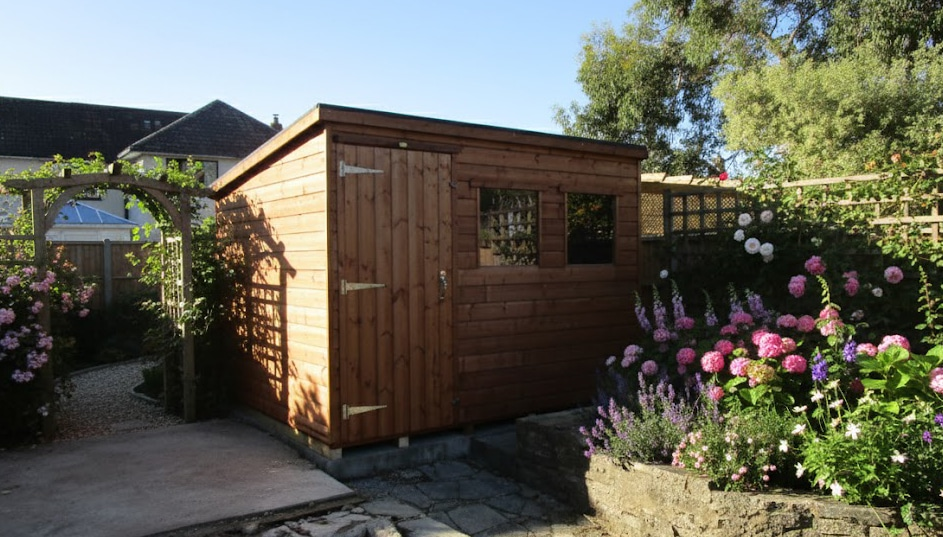 Wooden garden shed with floral border, surrounded by blooming pink flowers and green foliage in sunny garden setting